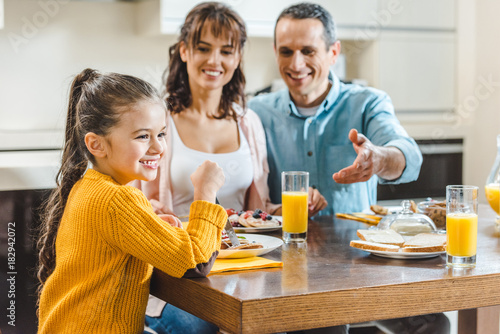 cheerful family sitting at table with pancakes, father pointing on juice by hand at kitchen