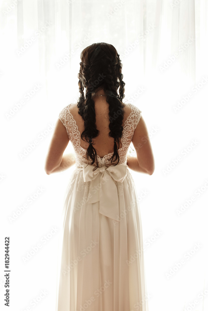 Look from behind at pretty bride in long dress standing before a bright window