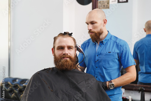 Male professional hairdresser serving client by clipper. Ginger handsome brutal stylish young man with thick big beard and short hair getting trendy haircut in black cape. Light white barber shop room