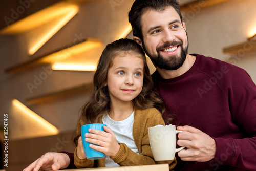 smiling father and daughter holding cups of cacao with marshmallow and looking away