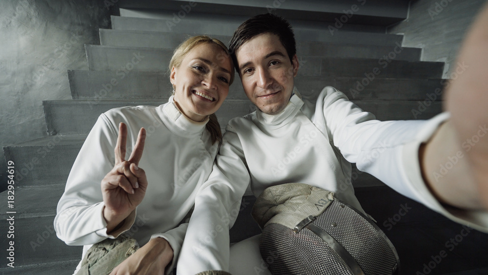 Fototapeta premium POV of Two young smiling fencers man and woman taking selfie on smartphone camera after fencing training indoors