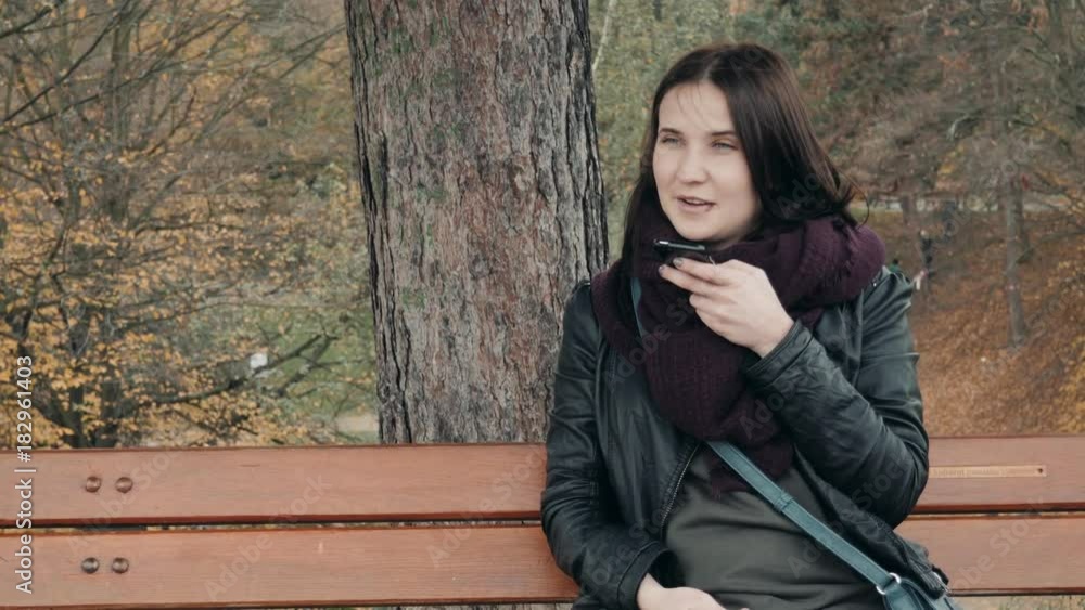 Young Smiling Woman Using Smartphone Sitting On Bench In Park. Beautiful European Girl Making Voice Message On Phone Young Smiling Woman Using Smartphone Sitting On Bench In Park. Beautiful European Girl Making Voice Message On Phone