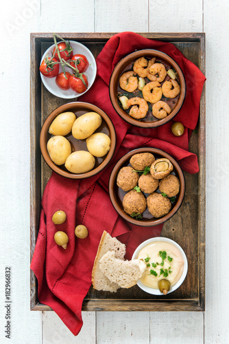 spanish tapas finger food, baked olives, shrimps, potatoes, tomato and garlic dip, party appetizers on a dark wooden tray with a red napkin on white painted wood, flat top view from above