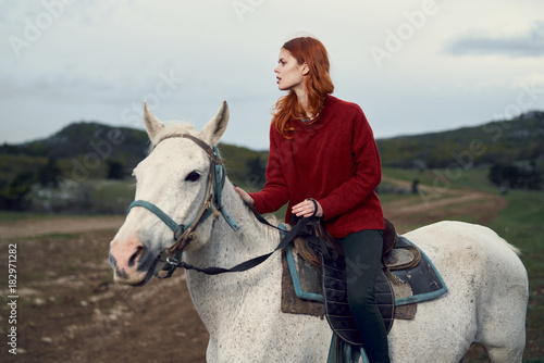 Young beautiful woman in a red sweater riding a horse on a white horse in the mountains, nature
