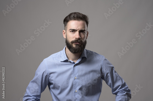 Young serious upset bearded business man with arms on hips and intense look at camera against gray studio background. 