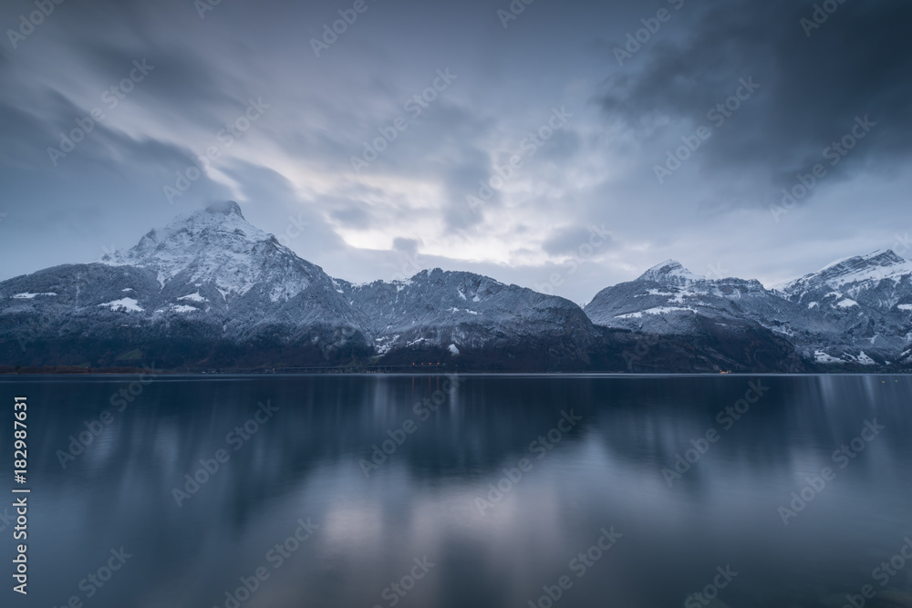 Mountains in overcast. Winter panorama. The mountains are reflected in the mirror water of the lake. Long exposure. Overcast. European Switzerland.
