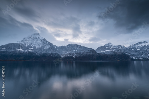 Mountains in overcast. Winter panorama. The mountains are reflected in the mirror water of the lake. Long exposure. Overcast. European Switzerland.