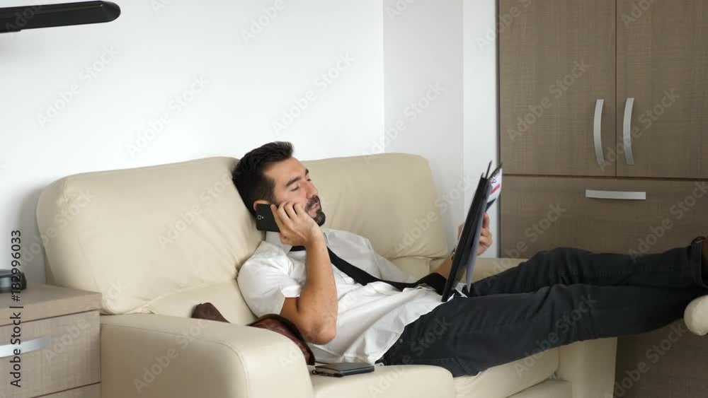 Revealing shot of tired businessman on the couch talking on the phone and examing some documents and charts Revealing shot of tired businessman on the couch talking on the phone and examing some documents and charts
