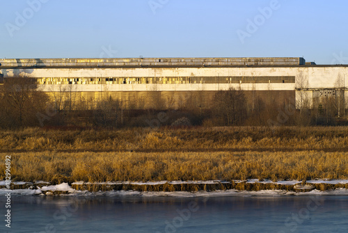 winter snowless landscape: some industrial building on the overgrown with dry grass and bushes bank of the canal, in the setting sun
