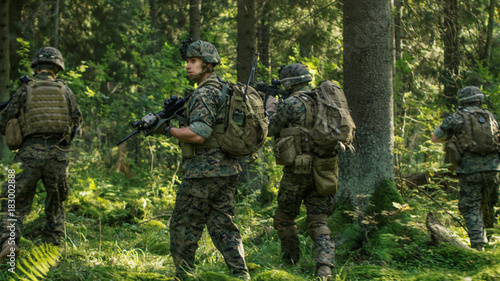 Squad of Fully Equipped Soldiers in Camouflage on a Reconnaissance Military Mission, Rifles in Firing Position. They're Moving in Formation Through Dense Forest. Back View Shot.