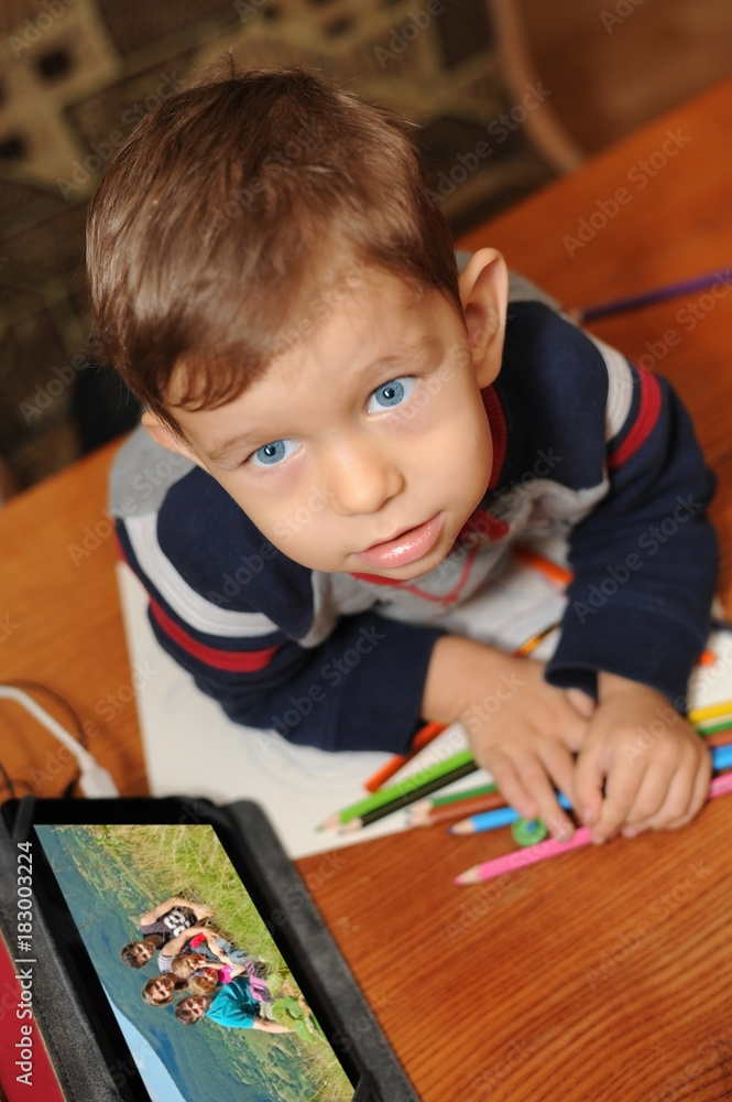 A little boy three years old paints with colored pencils on paper and watch family photos on a tablet A little boy three years old paints with colored pencils on paper and watch family photos on a tablet