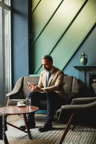 Middle-aged businessman drinking coffee and uses mobile phone in the loby