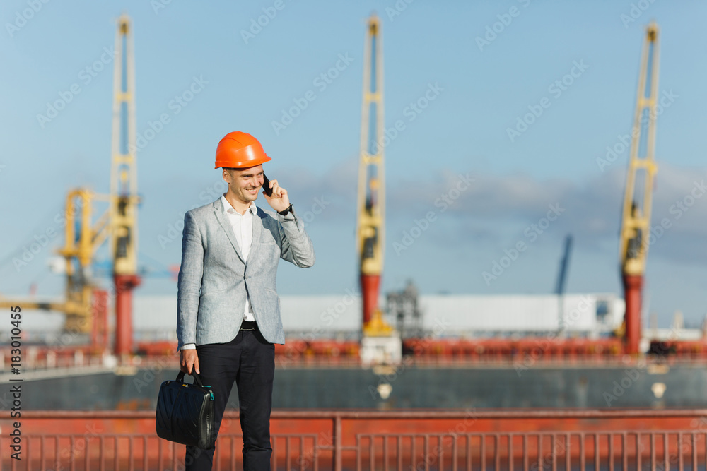 Handsome young unshaven successful business man in gray suit and protective construction orange helmet holding case, talking on mobile phone, walking in sea port against cargo ship, crane background Handsome young unshaven successful business man in gray suit and protective construction orange helmet holding case, talking on mobile phone, walking in sea port against cargo ship, crane background