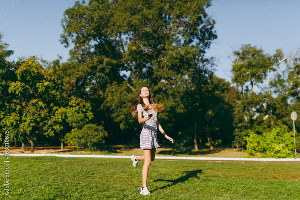 Young pretty girl with long brown hair dressed in light clothes staying on green lawn grass in the park on trees background. Summer sunny time. Young pretty girl with long brown hair dressed in light clothes staying on green lawn grass in the park on trees background. Summer sunny time.