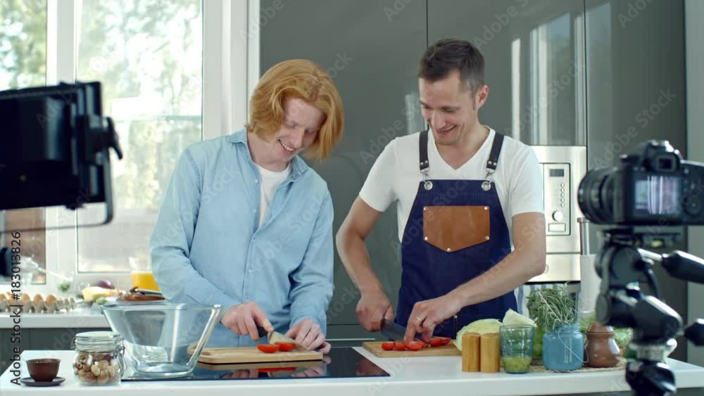 Medium shot of professional chef in apron and young man with ginger hair slicing tomatoes and chatting during filming of cooking show in modern kitchen Medium shot of professional chef in apron and young man with ginger hair slicing tomatoes and chatting during filming of cooking show in modern kitchen