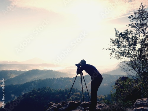 Nature photographer stay at tripod on summit and thinking. Hilly foggy landscape