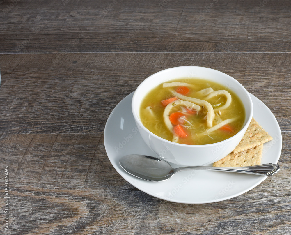 Overhead view of homemade chicken noodle soup with spoon and whole grain saltine crackers on rustic weathered wood background. Ingredients noodles, carrots, celery, chicken, broth. Overhead view of homemade chicken noodle soup with spoon and whole grain saltine crackers on rustic weathered wood background. Ingredients noodles, carrots, celery, chicken, broth.
