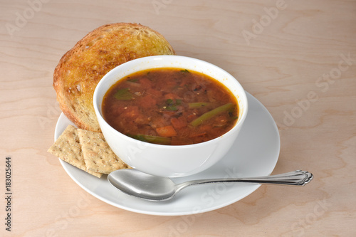Bowl of hot garden vegetable soup, spoon, whole grain saltine crackers, and sour dough toast on wooden table