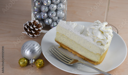 Isolated vanilla cheescake with a layer of whipped cream, fork,  and wine glass stem on a hickory wood table.  Christmas ornaments in the background.