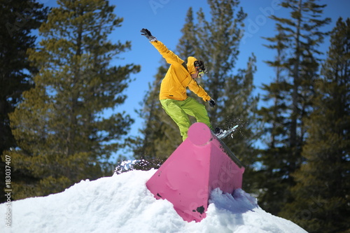 Young men jib on a snowboard in terrain park wearing yellow jacket and green pants. It is snow time during winter in a mountain ski resort in California.