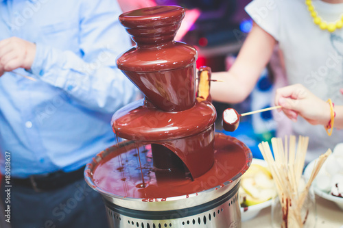 Vibrant Picture of Chocolate Fountain Fontain on a children kids birthday party with a kids playing around and dipping marshmallows and fruits into the fountain
