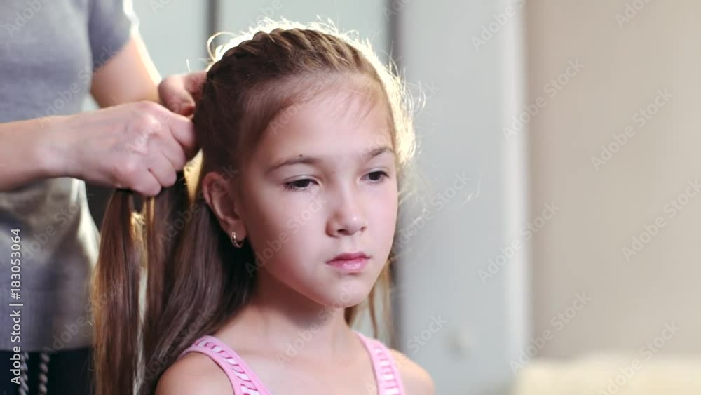 An attractive woman does her hairstyle for her little daughter. A woman is weaving a pigtail for her daughter.