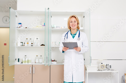 Confident pharmacist in whitecoat standing by cabinet with variety of bottle with medication