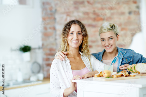 Happy girlfriends in casualwear sitting by table in the kitchen at home