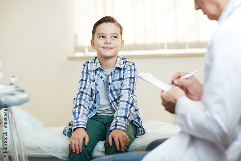 Happy little boy looking at his doctor while he making prescriptions and notes in medical form Happy little boy looking at his doctor while he making prescriptions and notes in medical form