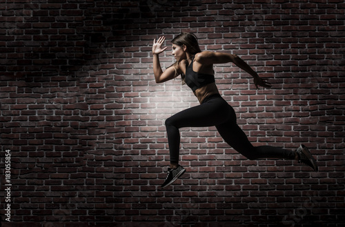 Beautiful muscular fitness model wearing long black tights and black sports bra showing her six pack abs doing a running leap or jump with a dramatic lit red brick background  © Paul