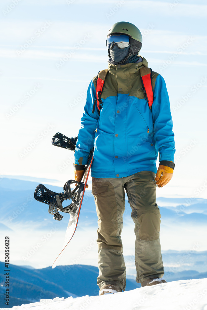 Young snowboarder standing at the top of a mountain and holding his snowboard with one hand