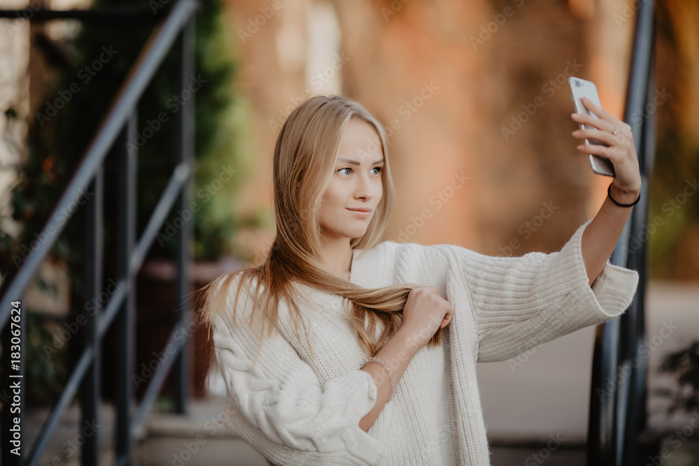 Beautiful casual blonde stylish fashion woman with a phone in her hand make selfie. European buildings and street on a background. Copy space Beautiful casual blonde stylish fashion woman with a phone in her hand make selfie. European buildings and street on a background. Copy space