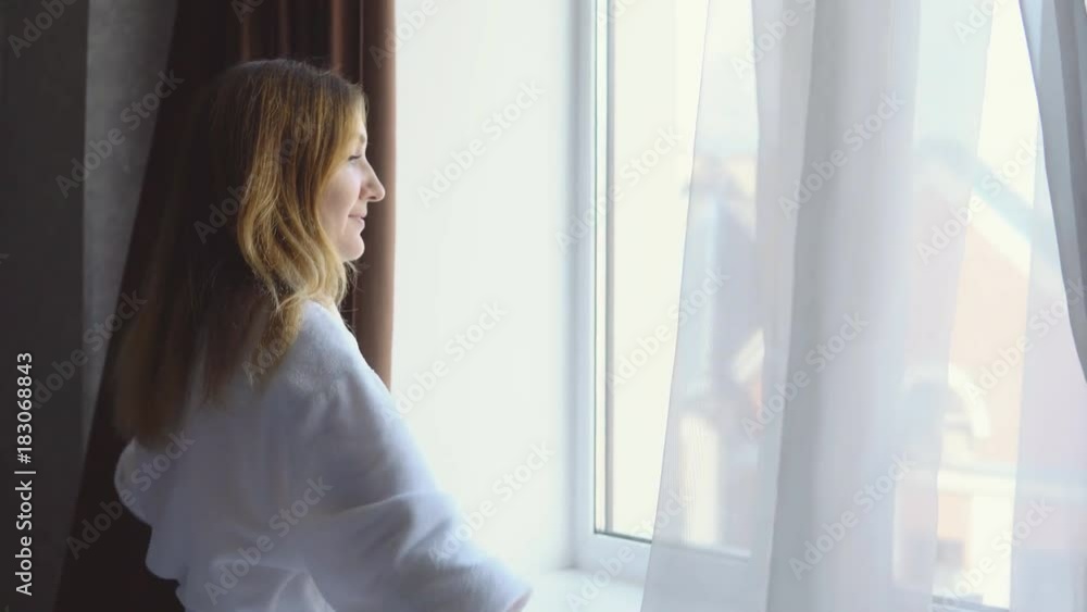 Attractive young positive woman in white robe looks out the window in the early morning and smiles Attractive young positive woman in white robe looks out the window in the early morning and smiles