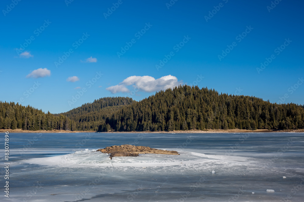 Winter landscape. Frozen lake, pine forest. Bulgaria, Rhodopes mountains, Shiroka Polyana lake. Rocky island in front.