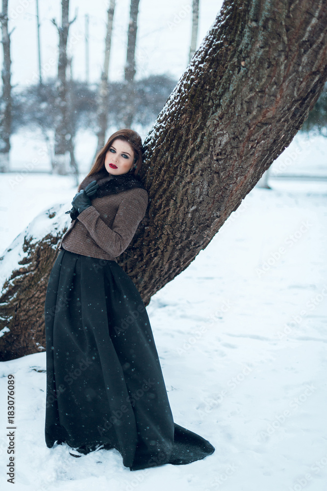Portrait of a beautiful young woman with red lips and smoky eyes, dressed in a coat, a long black skirt and a hat, in a snowy park Portrait of a beautiful young woman with red lips and smoky eyes, dressed in a coat, a long black skirt and a hat, in a snowy park