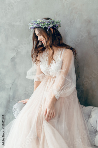 A girl in a beige peignoir, with a wreath of flowers on her head, poses in the studio loft, fine art wedding style photo