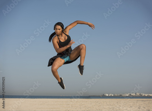 Beautiful Inter-racial Black Asian Fitness Model does an explosive dance moves wearing headphones on a white sandy beach on a clear blue sunny morning. 
