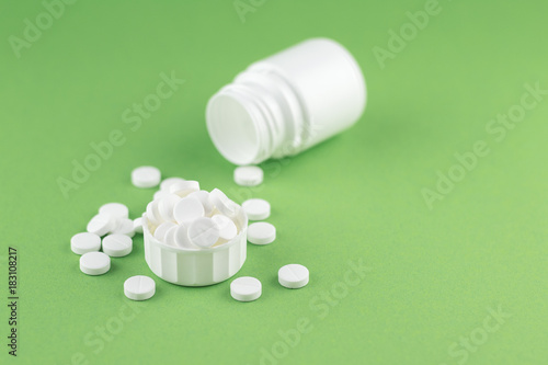 Close up white pill bottle with spilled out pills and capsules in cap on pistachio green background with copy space. Focus on foreground, soft bokeh. Pharmacy drugstore concept