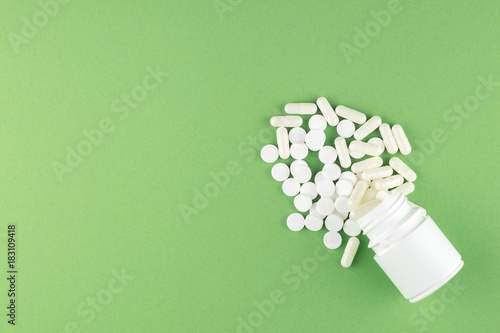 Close up white pill bottle with spilled out pills and capsules on pistachio green background with copy space. Focus on foreground, soft bokeh. Pharmacy drugstore concept. Top view