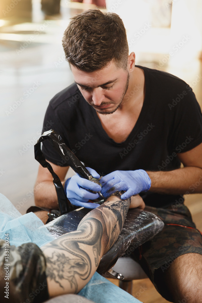 Professional tattooer artist doing picture on hand of man by machine black ink from a jar. Tattoo art on body. Equipment for making tattoo art. Master makes tattooed in light studio. Professional tattooer artist doing picture on hand of man by machine black ink from a jar. Tattoo art on body. Equipment for making tattoo art. Master makes tattooed in light studio.
