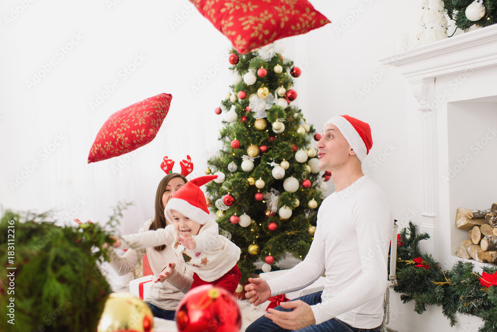Happy young cheerful parents fighting pillows with cute little son. Child boy in red hat in light room at home with decorated New Year tree. Christmas good mood. Family, love and holiday 2018 concept.