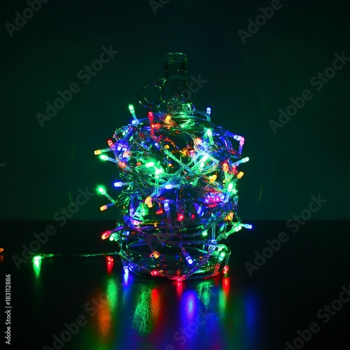 A colorful electric glowing Christmas garland wire wrapped around the glass bottle.