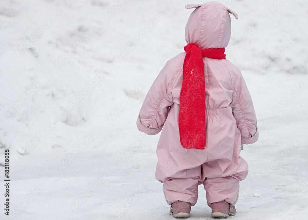 Small child in pink overalls with bunny ears lies and the bright scarf is walking on the snow in winter. Full body portrait of kid from behind against snow cover background.