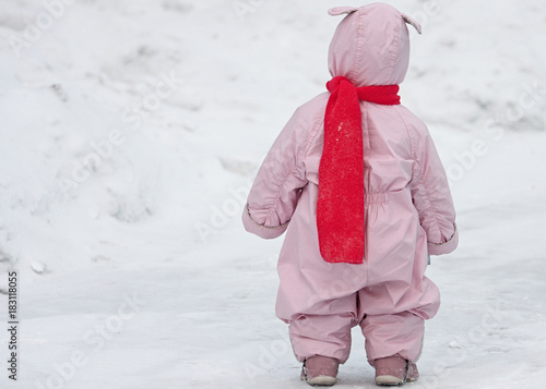 Obraz na płótnie Small child in pink overalls with bunny ears lies and the bright scarf is walking on the snow in winter. Full body portrait of kid from behind against snow cover background. w salonie