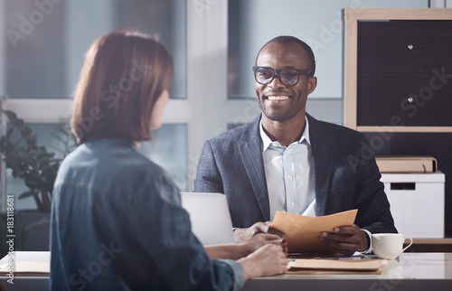 Glad to hear it. Portrait of optimistic young qualified african manager is looking at his colleague female with smile while sitting at table and having pleasant communication. Back view of woman photo