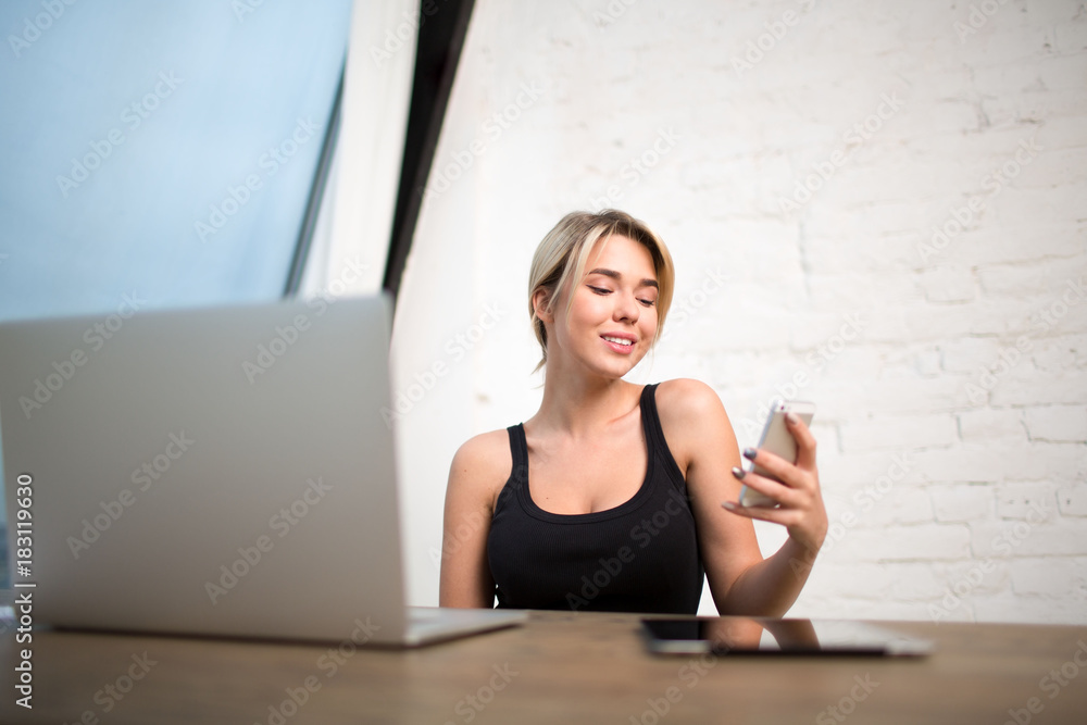 Cheerful female publication specialist reading private information on mobile phone, sitting at work table with open laptop computer. Smiling businesswoman having on-line video call on cell telephone