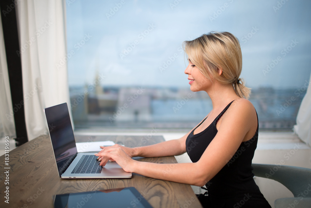 Naklejka premium Smiling woman publication specialist typing text on modern laptop computer, sitting at the table in office. Pretty female magazine editor keyboarding on net-book. Financier using notebook for chat