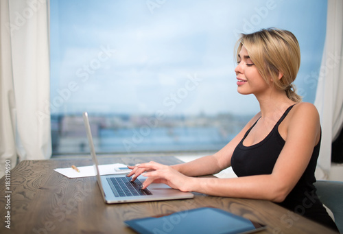 Smiling woman office worker keyboarding on modern laptop computer, sitting at table near window. Cheerful female secretary typing on portable net-book. Workstation with gadgets. Student using notebook