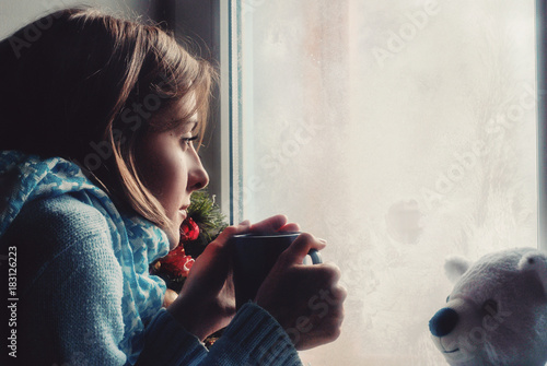 Sad upset lonly beautiful young woman with the cup in sweater dreaming near the frozen window at home photo
