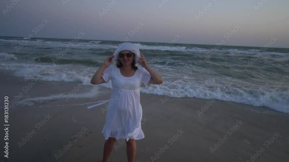 Young beautiful woman is standing by the sea at sunset. In a white dress and a white hat. She looks at the camera and smiles. The woman is pregnant. Young beautiful woman is standing by the sea at sunset. In a white dress and a white hat. She looks at the camera and smiles. The woman is pregnant.